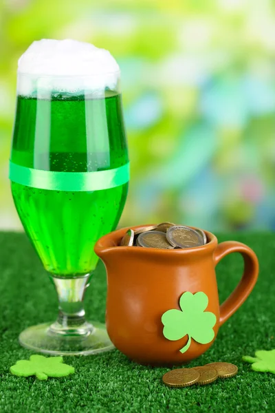 Glass of green beer and pitcher with coins on grass on natural background — Stock Photo, Image