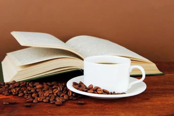 Чашка кофе с кофейными зёрнами и книга на деревянном столе на коричневом фоне Лицензионные Стоковые Фото