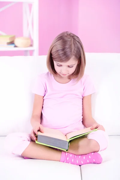 Malá holčička, čtení knihy sedí na pohovce v místnosti — Stock fotografie