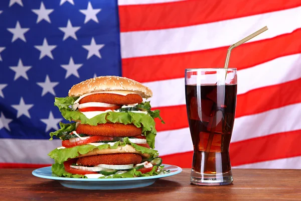 Огромный гамбургер на цветной тарелке и стакане холодного напитка, на фоне флага США — стоковое фото