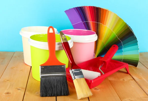 Paint pots, paintbrushes and coloured swatches on wooden table on blue background — Stock Photo, Image