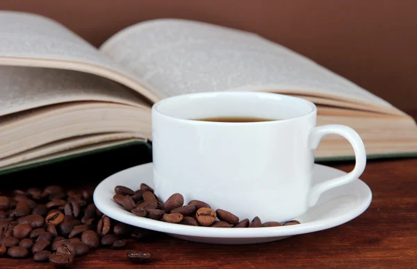 Kopp kaffe med kaffebönor och bok om träbord på brun bakgrund Stockfoto