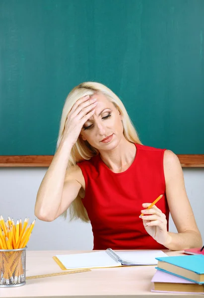 Tired teacher sitting at table on blackboard background — Stock Photo, Image