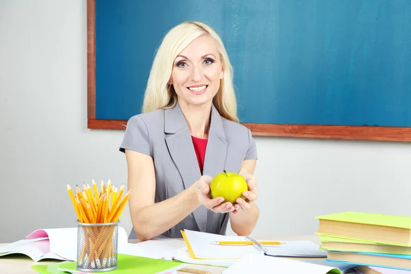 Schoolleraar met apple zittend aan tafel op blackboard achtergrond — Stockfoto