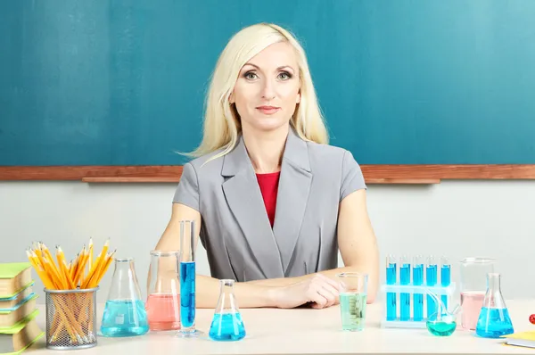 Chemistry teacher sitting at table on blackboard background — Stock Photo, Image