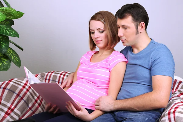 Jovem grávida segurando livro e seu marido no sofá em casa — Fotografia de Stock