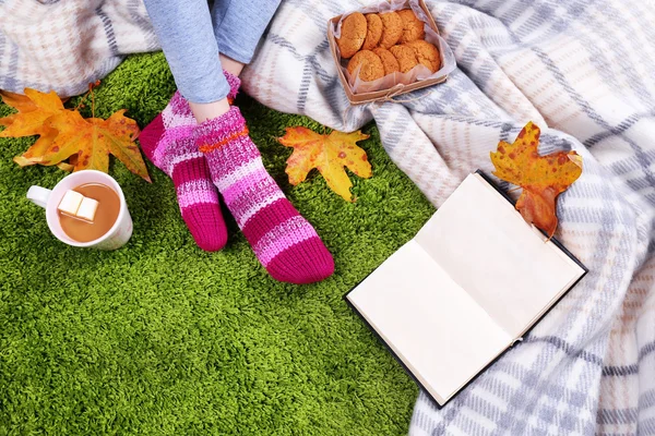 Composición con cuadros calientes, libro, taza de bebida caliente y piernas femeninas, sobre fondo de alfombra de color — Foto de Stock