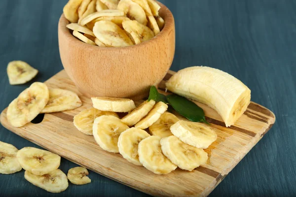 Frische und getrocknete Bananenscheiben in Schüssel, auf Schneidebrett, auf Holzgrund — Stockfoto