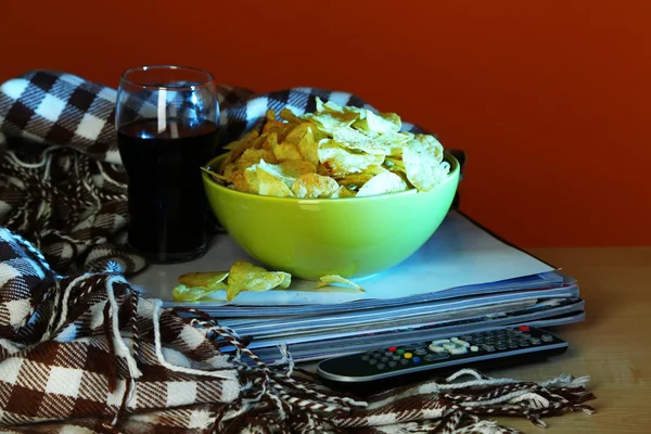 Chips dans le bol, le cola et la télévision à distance sur table en bois sur fond coloré — Photo