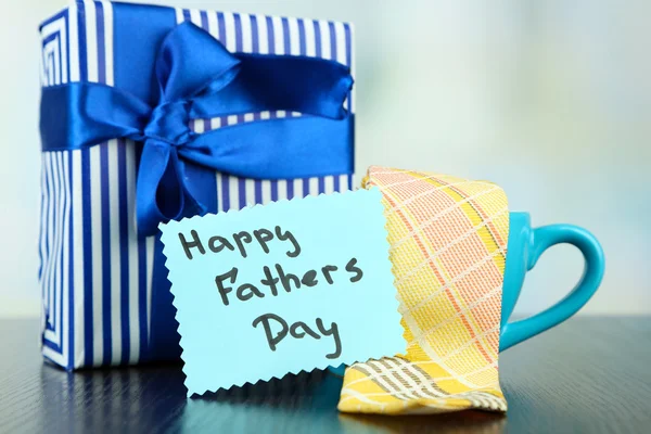 Тег Happy Fathers Day с подарочными коробками, чашкой и галстуком, на деревянном столе, на светлом фоне — стоковое фото