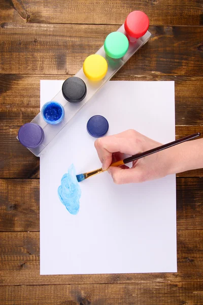 Hand som håller penseln med färger och papper på trä bakgrund — Stockfoto