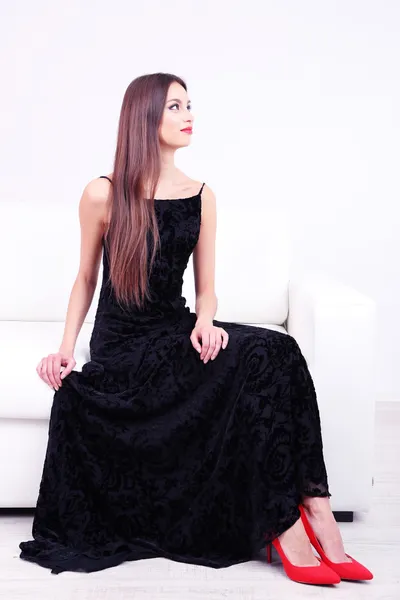 Mooie jonge vrouw in zwarte jurk op sofa op witte achtergrond — Stockfoto