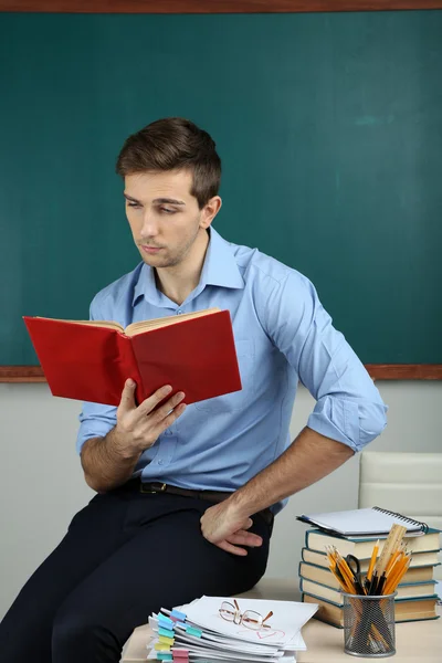 Junge Lehrerin sitzt mit Buch auf Schreibtisch im Klassenzimmer — Stockfoto