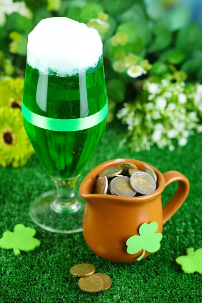 Glass of green beer and pitcher with coins on grass close-up — Stock Photo, Image