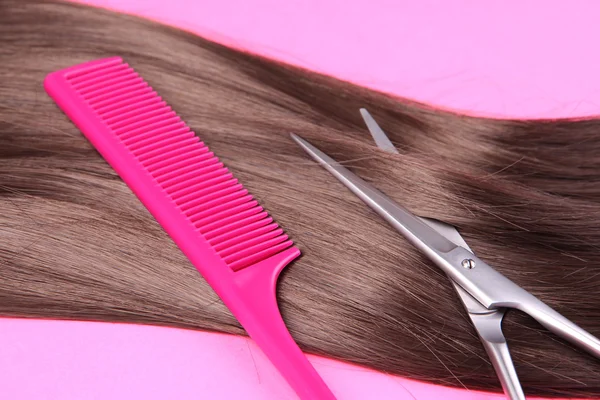 Длинные каштановые волосы с расческой и ножницами на розовом фоне — стоковое фото