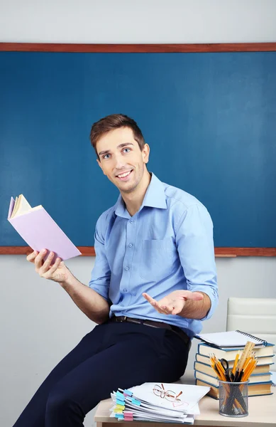 Junge Lehrerin sitzt mit Buch auf Schreibtisch im Klassenzimmer — Stockfoto