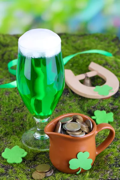 Glass of green beer, pitcher with coins and horseshoe on grass close-up — Stock Photo, Image