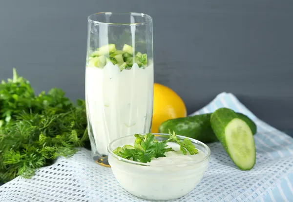 Gurkenjoghurt in Glas und Schüssel, auf Farbserviette, auf Holztisch, auf grauem Hintergrund — Stockfoto