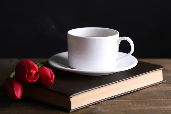 暗い背景上のテーブルの上に花と本にホット コーヒーのカップ — ストック写真