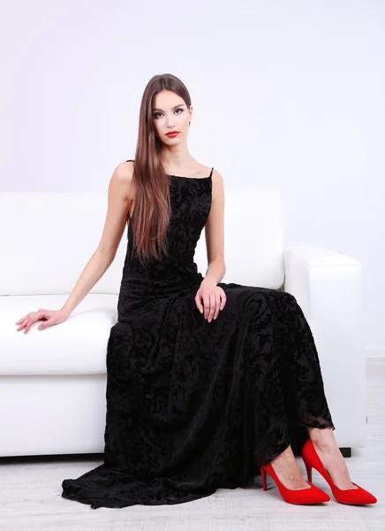 Mooie jonge vrouw in zwarte jurk op sofa op witte achtergrond — Stockfoto