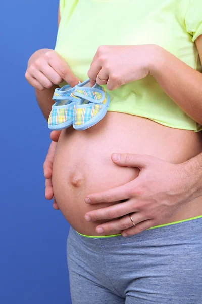 Pregnant woman holding baby shoes with her husband on blue background — Stock Photo, Image