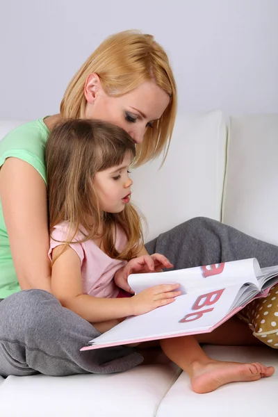 Jolie petite fille livre de lecture avec mère sur canapé sur fond gris — Photo