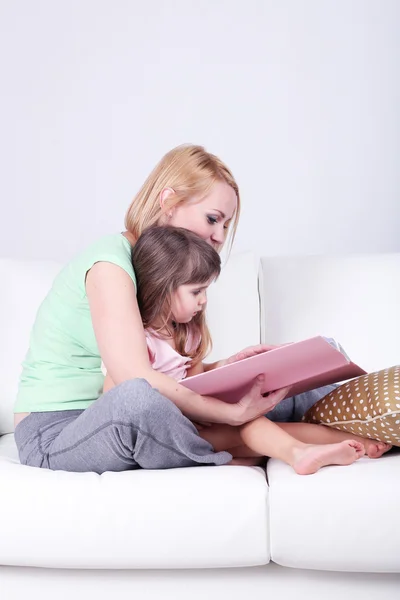 漂亮的小女孩和妈妈坐在沙发上灰色的背景上看书 — 图库照片