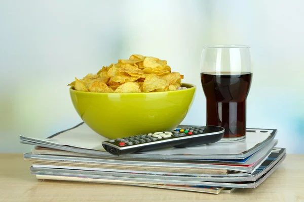 Čipy v misce, cola a tv ovladač na dřevěný stůl na pozadí místnosti — Stock fotografie