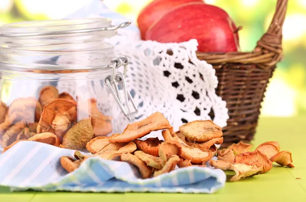 Composition with dried apples in glass jar and fresh apples in basket, on napkin, on bright background — Stock Photo, Image