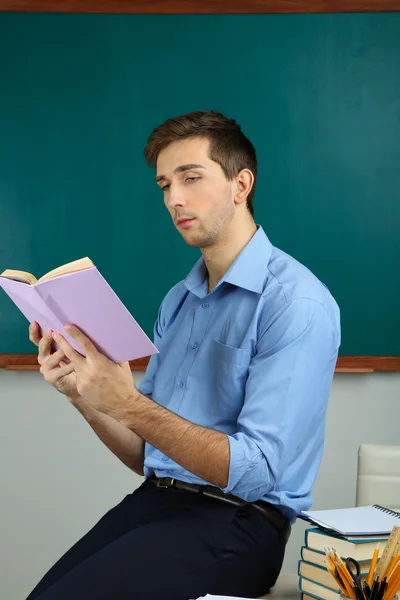 Junge Lehrerin sitzt mit Buch auf Schreibtisch im Klassenzimmer — Stockfoto