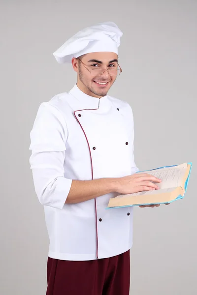 Professionele chef-kok in het witte uniform en hoed, bedrijf van boek, op grijze achtergrond — Stockfoto