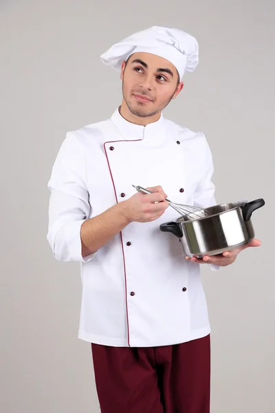 Chef profesional en uniforme blanco y sombrero, sosteniendo la sartén en sus manos, sobre fondo gris — Foto de Stock