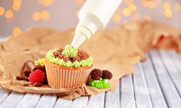 Confectioner decorating tasty cupcake with butter cream, on color wooden table, on lights background — Stock Photo, Image