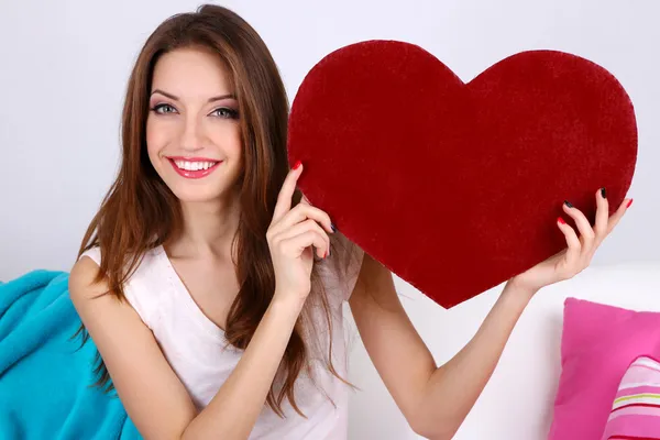 Hermosa joven sentada en un sofá con corazón decorativo sobre fondo gris —  Fotos de Stock