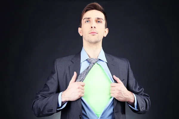 Young business man tearing apart his shirt revealing superhero suit, on dark background — Stock Photo, Image