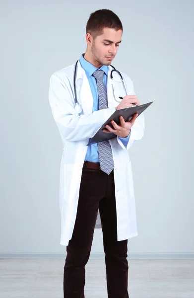 Médico masculino de pé com pasta, em fundo cinza — Fotografia de Stock