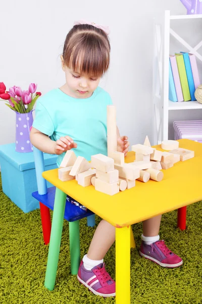 Little girl plays with construction blocks sitting at table in room — Stock Photo, Image