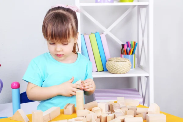 Little girl plays with construction blocks sitting at table in room — Stock Photo, Image