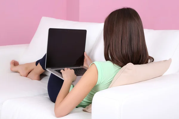 Young woman sitting on sofa with laptop on pink background — Stock Photo, Image