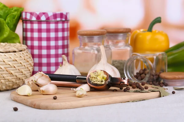 Samenstelling met knoflook pers, verse knoflook en glazen potten met specerijen op houten tafel, op lichte achtergrond — Stockfoto