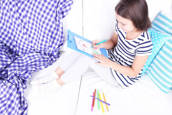 Beautiful little girl sitting on sofa and drawing picture, on home interior background — Stock Photo, Image