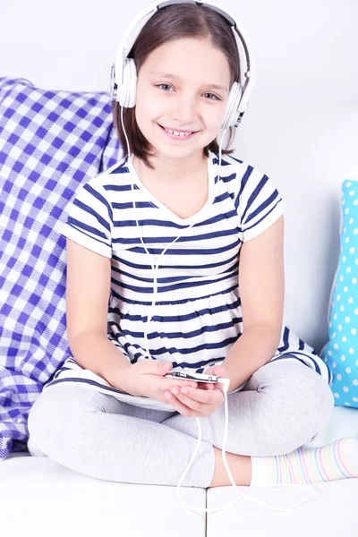 Beautiful little girl sitting on sofa and listening to music, on home interior background — Stock Photo, Image