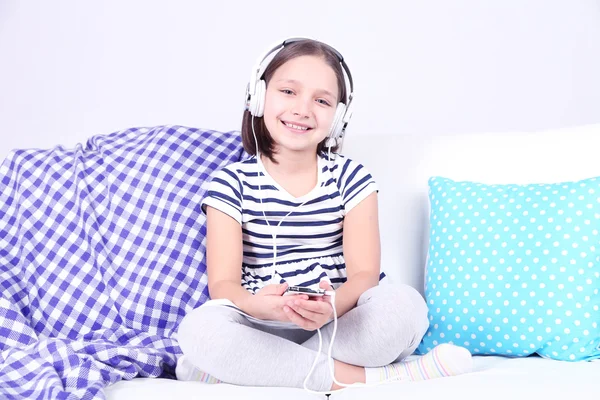 Beautiful little girl sitting on sofa and listening to music, on home interior background — Stock Photo, Image