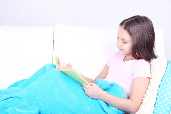 Menina bonita sentada no sofá com livro, em casa fundo interior — Fotografia de Stock