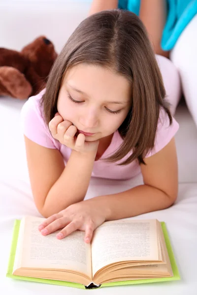 Hermosa niña sentada en el sofá con libro, en el fondo interior de casa — Foto de Stock