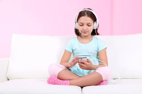 Beautiful little girl sitting on sofa and listening to music, on home interior background — Stock Photo, Image