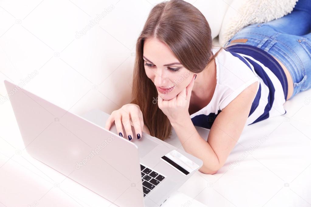 Young woman sitting with laptop on sofa and holding credit card in her ...