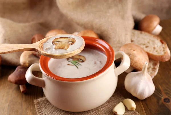 Sopa de cogumelos em colher e panela de madeira, sobre mesa de madeira, sobre fundo de pano de saco — Fotografia de Stock