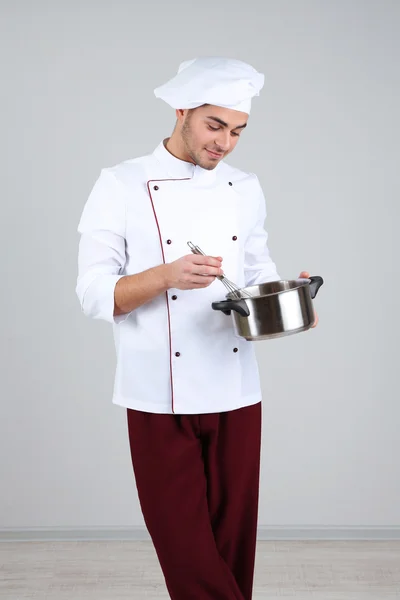 Професійний шеф-кухар в білій формі і капелюсі, тримає сковороду в руках, на сірому фоні — стокове фото