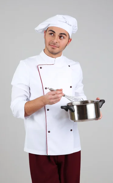 Chef profesional en uniforme blanco y sombrero, sosteniendo la sartén en sus manos, sobre fondo gris — Foto de Stock